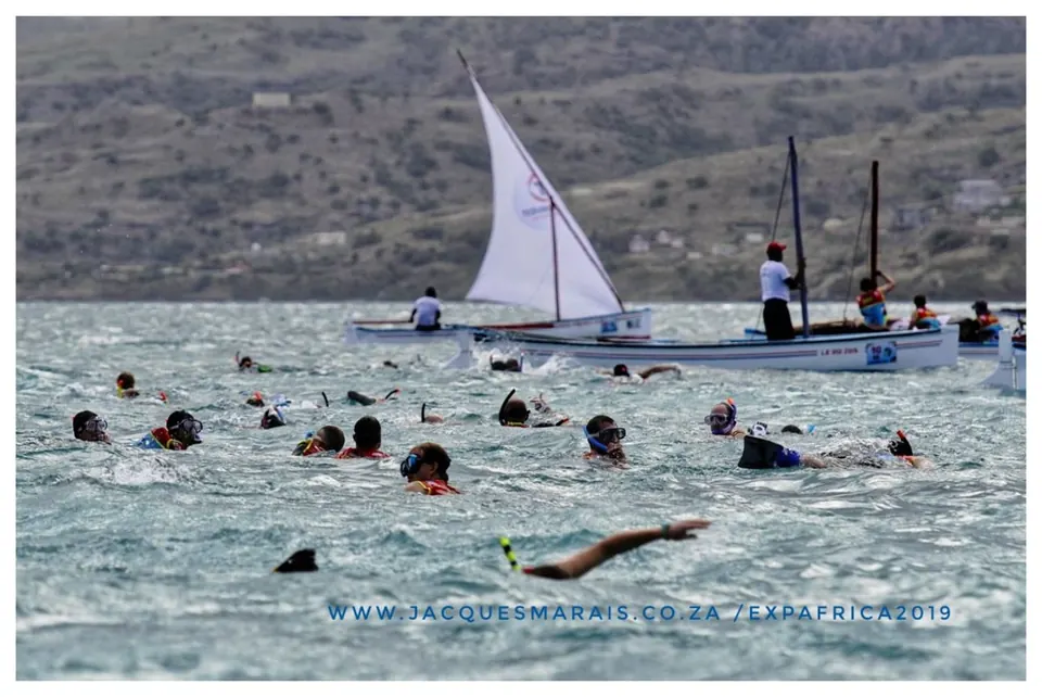 Swimmers in the sea beside traditional sailboats at the race start