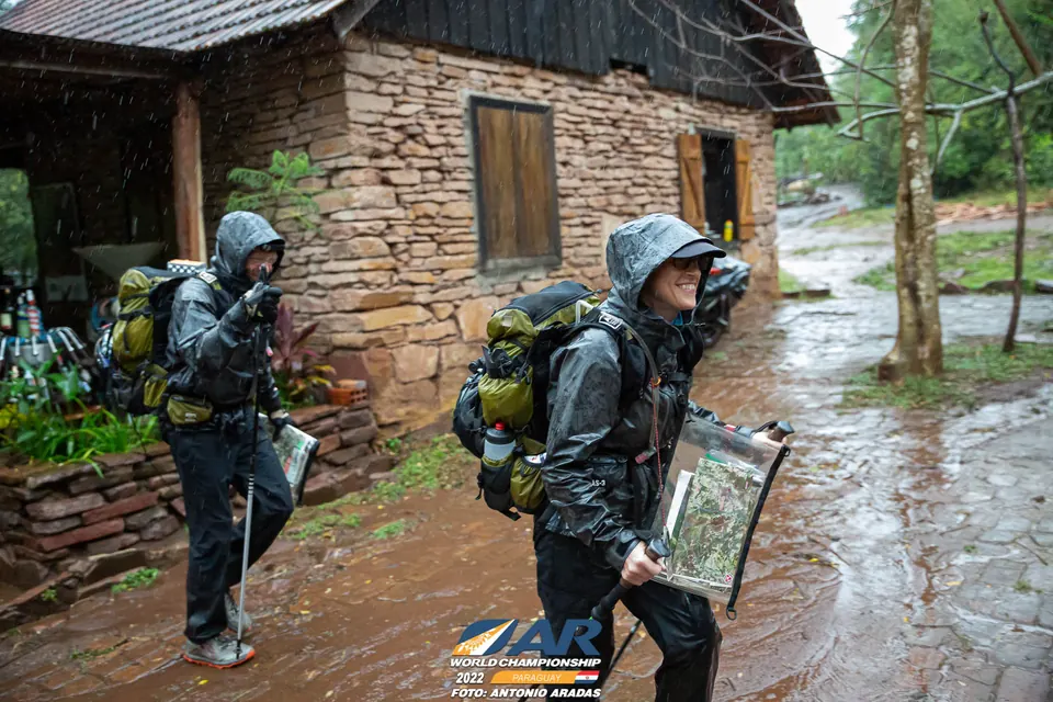 Racers in rain gear with packs and a map outside a stone building
