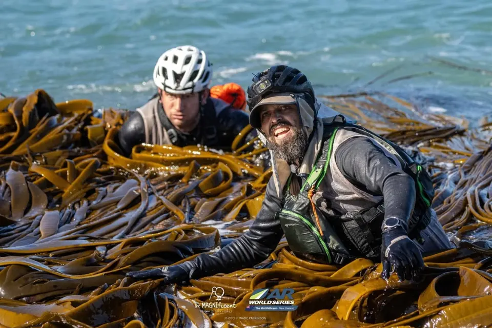 Two racers in helmets crawling through thick kelp on a shoreline