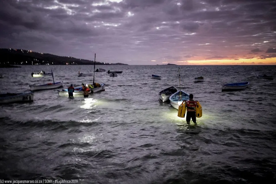 Racers wading through shallow water at dusk with headlamps and boats