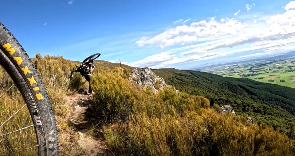 Mountain biker on a hillside trail, framed by a bike wheel in the foreground